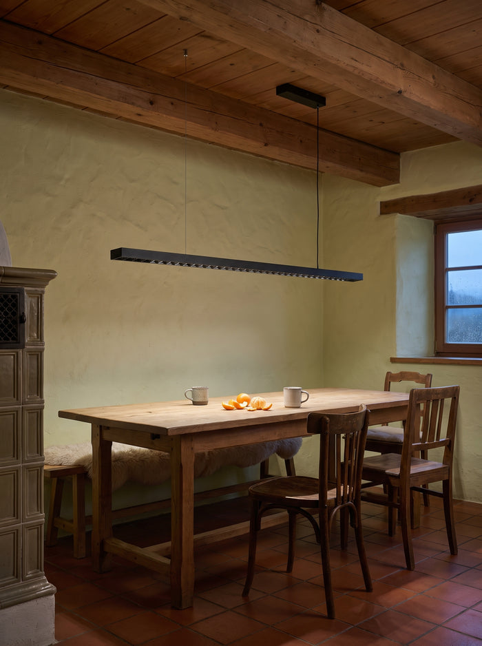 Schwarze lineare LED-Pendelleuchte über einem alten Eichentisch in einer Bauernstube mit Holzbalkendecke
