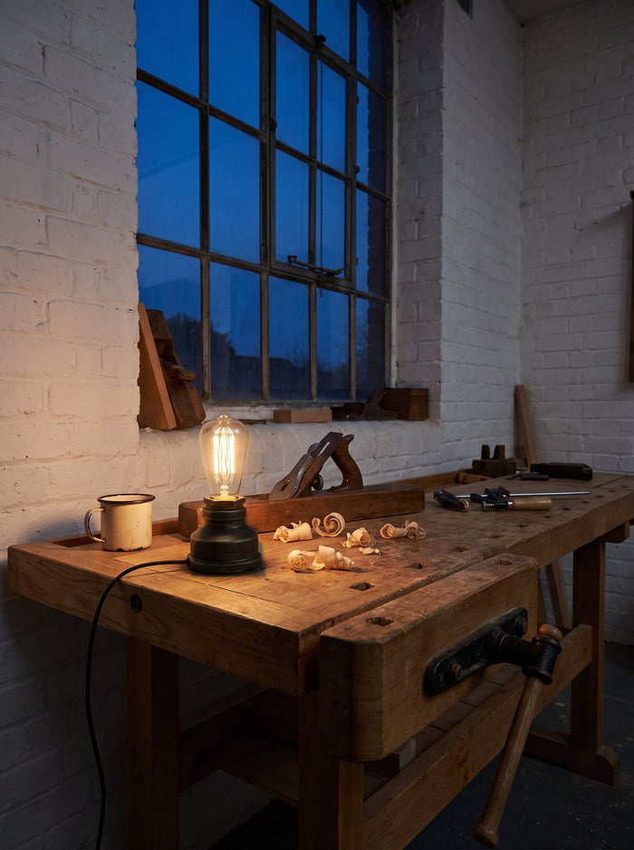 Industrie-Tischlampe im Wasserrohr-Design mit dunklem Gusssockel auf einer Hobelbank in einer Backsteinwerkstatt am Abend